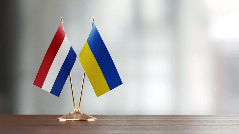 Dutch And Ukrainian Flag Pair On A Desk Over Defocused Background